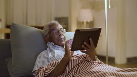 Elderly-black-man-lying-in-bed-surfing-net-using-digital-tablet