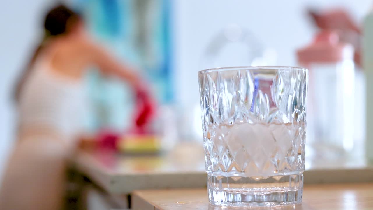 A woman cleans dishes in a sunlit kitchen, with a focus on a glass in the foreground