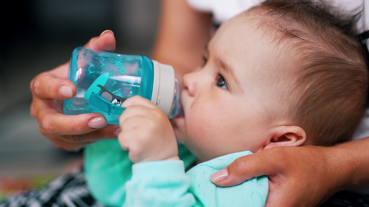 Nice baby boy drinking water from bottle. Careful mommy supporting her child and bottle from behind. Close up.
