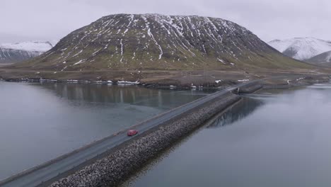 Vista-Aérea-Del-Coche-Rojo-Que-Se-Mueve-En-La-Carretera-Sobre-El-Lago-Glacial-En-El-Paisaje-De-Islandia