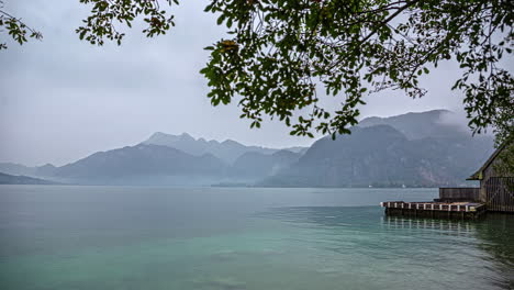 Vista-Al-Lago-Con-Niebla-Desplegada-Sobre-Majestuosas-Montañas,-Timelapse-De-La-Naturaleza-Bajo-La-Rama-De-Un-árbol