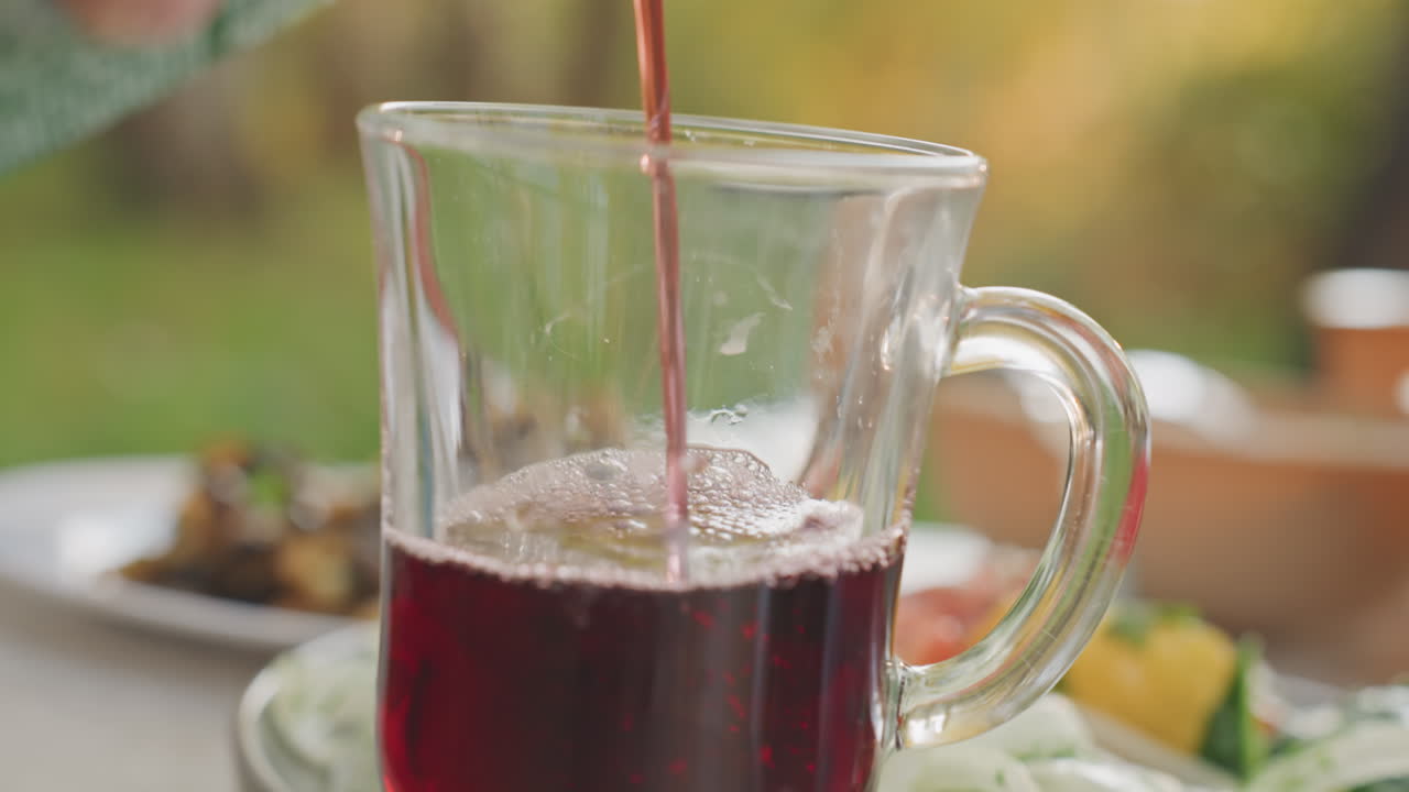 Bicchiere sul tavolo da campeggio che si riempie di rosso, primo piano di una cena serale con versamento da un cartone in una tazza, piatti e insalate al tenue bagliore di una lanterna, amici che si riuniscono intorno al tavolo, atmosfera autunnale accogliente.