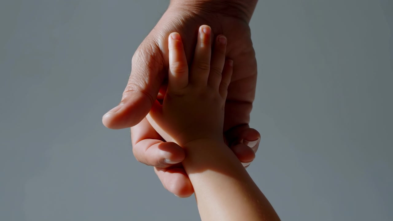 Centered close-up of an adult hand gently holding a toddler's hand against a smooth neutral backdrop. Soft directional light from the left highlights skin