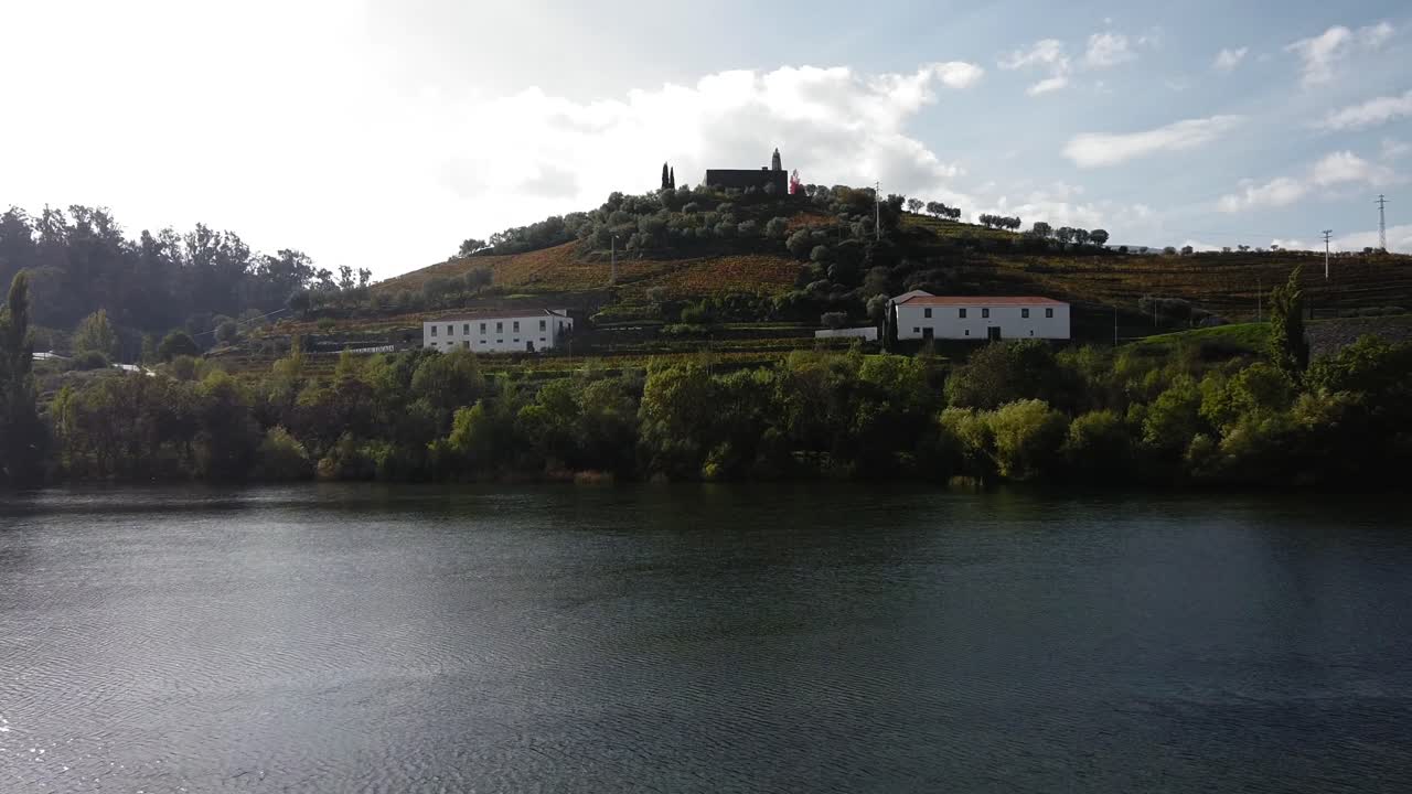 Riverside view of a picturesque Douro Valley landscape, featuring a wide river in the foreground and a terraced hillside with vineyards, traditional buildings, and a distant church under a cloudy sky.