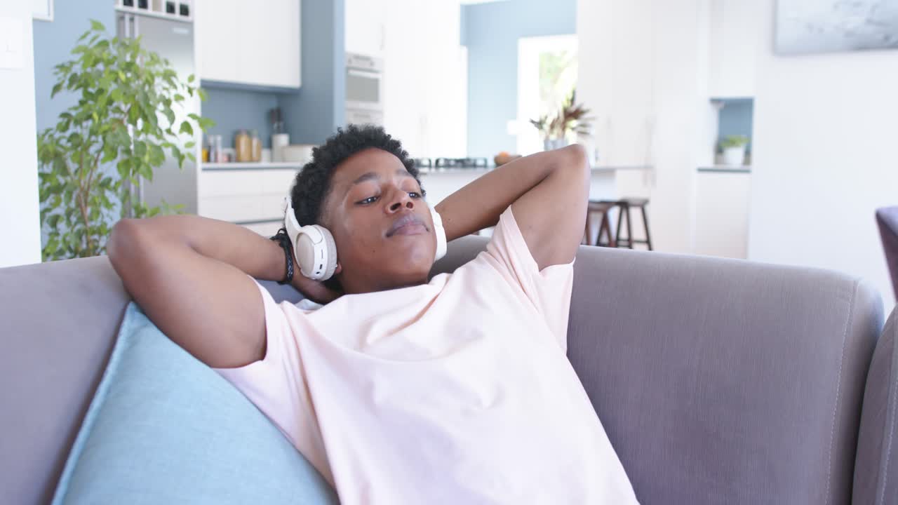 African American man lounging on gray sofa at home, listening with white headphones, relaxing