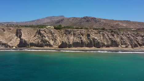 Aerial-flying-sideways-alongside-island,-rocky-cliff-and-blue-water-waves-in-the-Mediterranean-Sea-on-beach-in-Santorini,-Greece