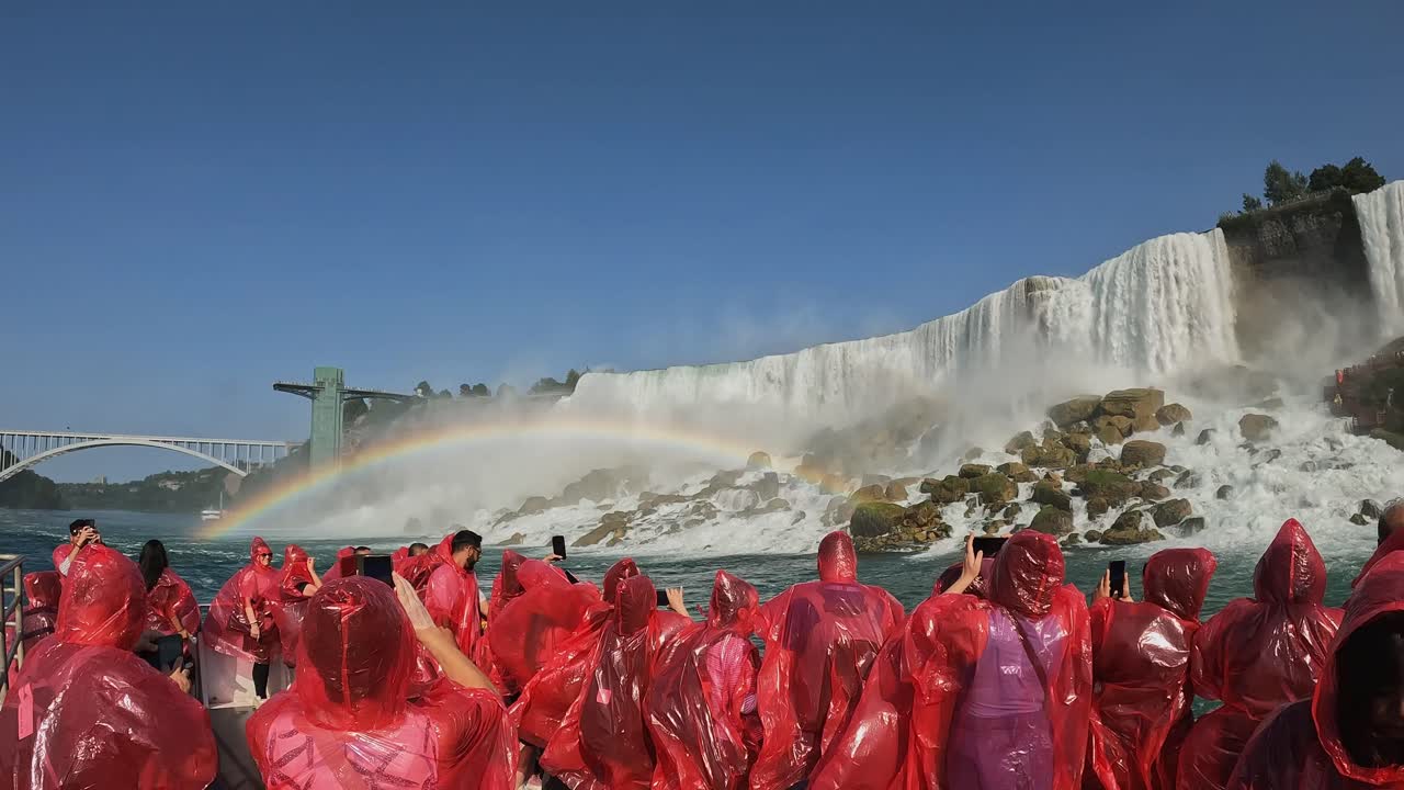 Group of tourists in red ponchos stands on a boat deck, admiring the majestic Niagara Falls. A vivid rainbow arcs over the mist, creating a dramatic scene on a clear, sunny day, Canada