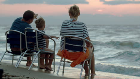 family of three sitting on chairs by sea at sunset