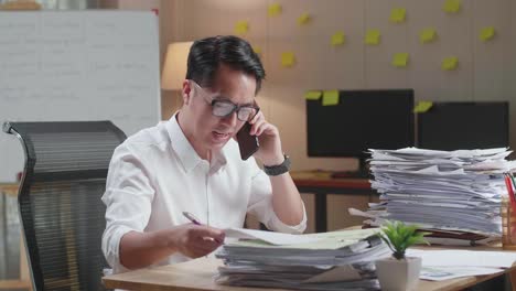 asian man talking on phone and taking note while working hard with documents at office