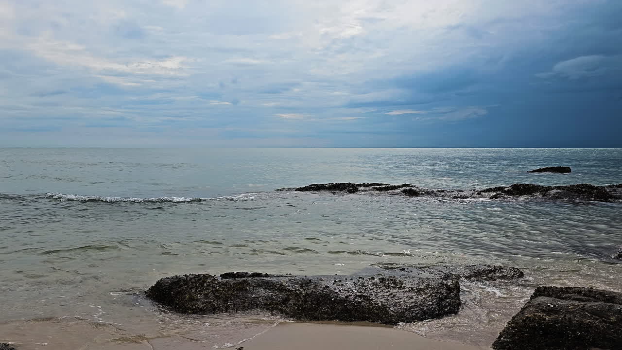 해변의 파도가 해가 지는 모래 해변에서 해안의 바위 위를 굴러서 폭우 구름과 함께 극적인 하늘을 흐르고 있습니다.
