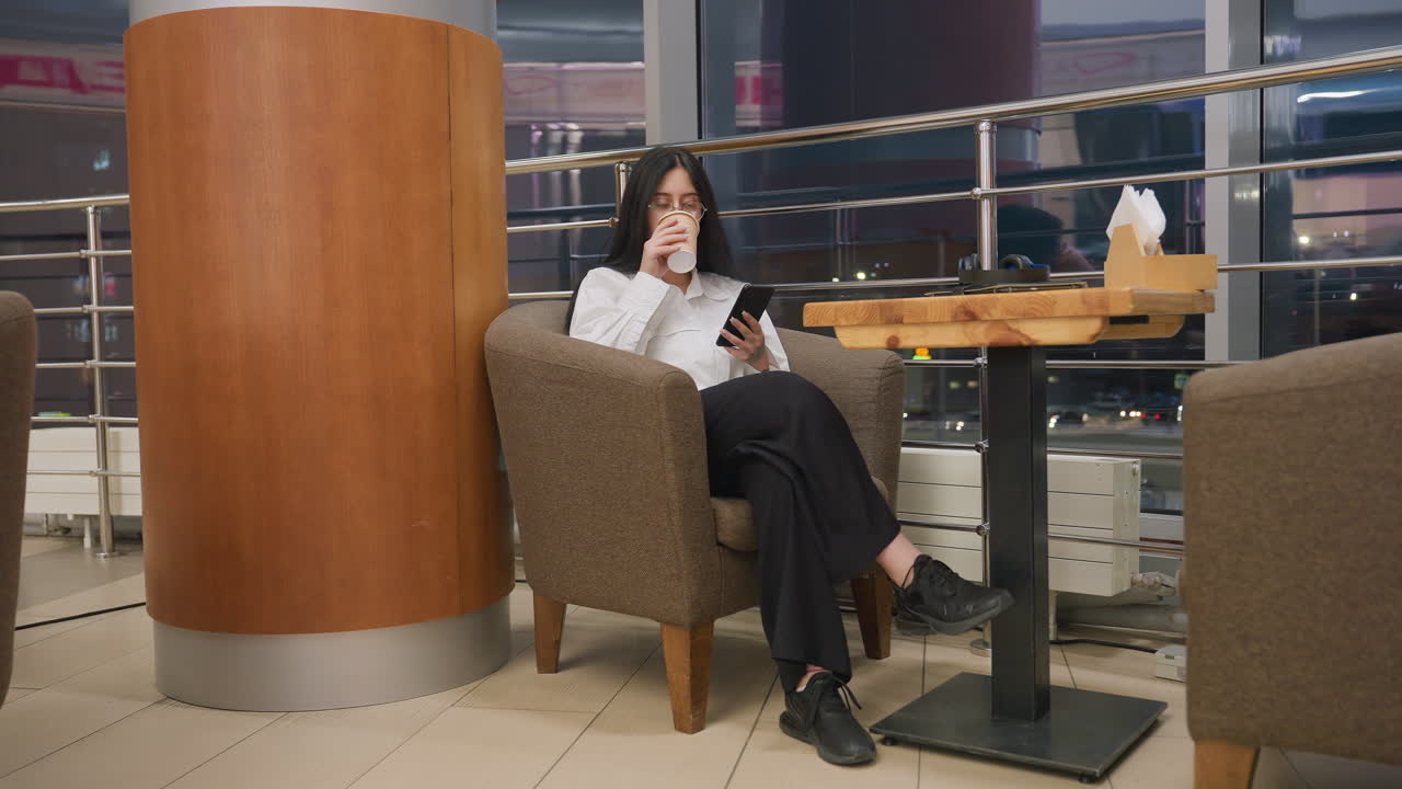 Woman sits comfortably in lounge chair sipping coffee and using phone while chatting indoors, table nearby holds tablet, headphones, and napkin holder, city lights glow through window in background