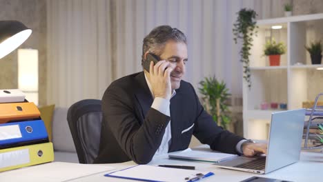 Happy-mature-businessman-working-on-laptop-while-talking-by-phone.