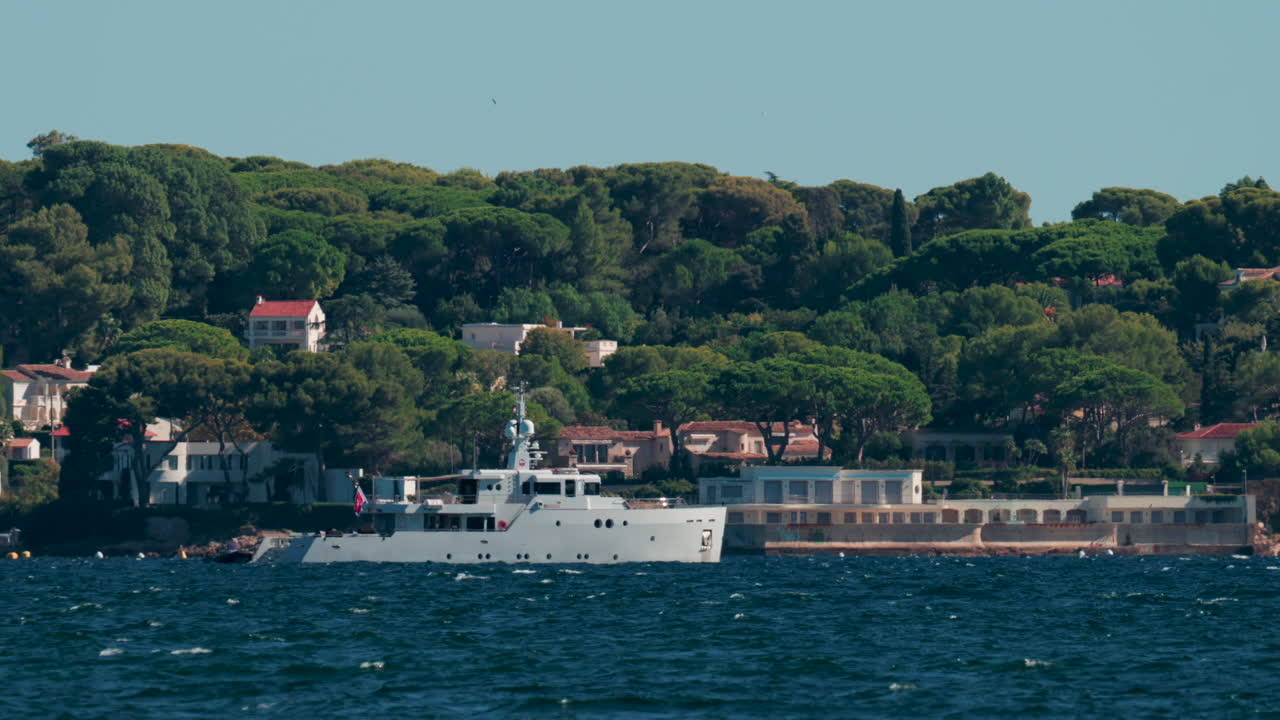 Luxury white yacht sailing near Mediterranean coastline with elegant villas surrounded by trees