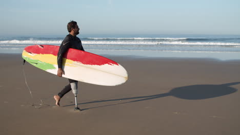 long shot of a male surfer with artificial leg walking along beach and holding surfboard under arm