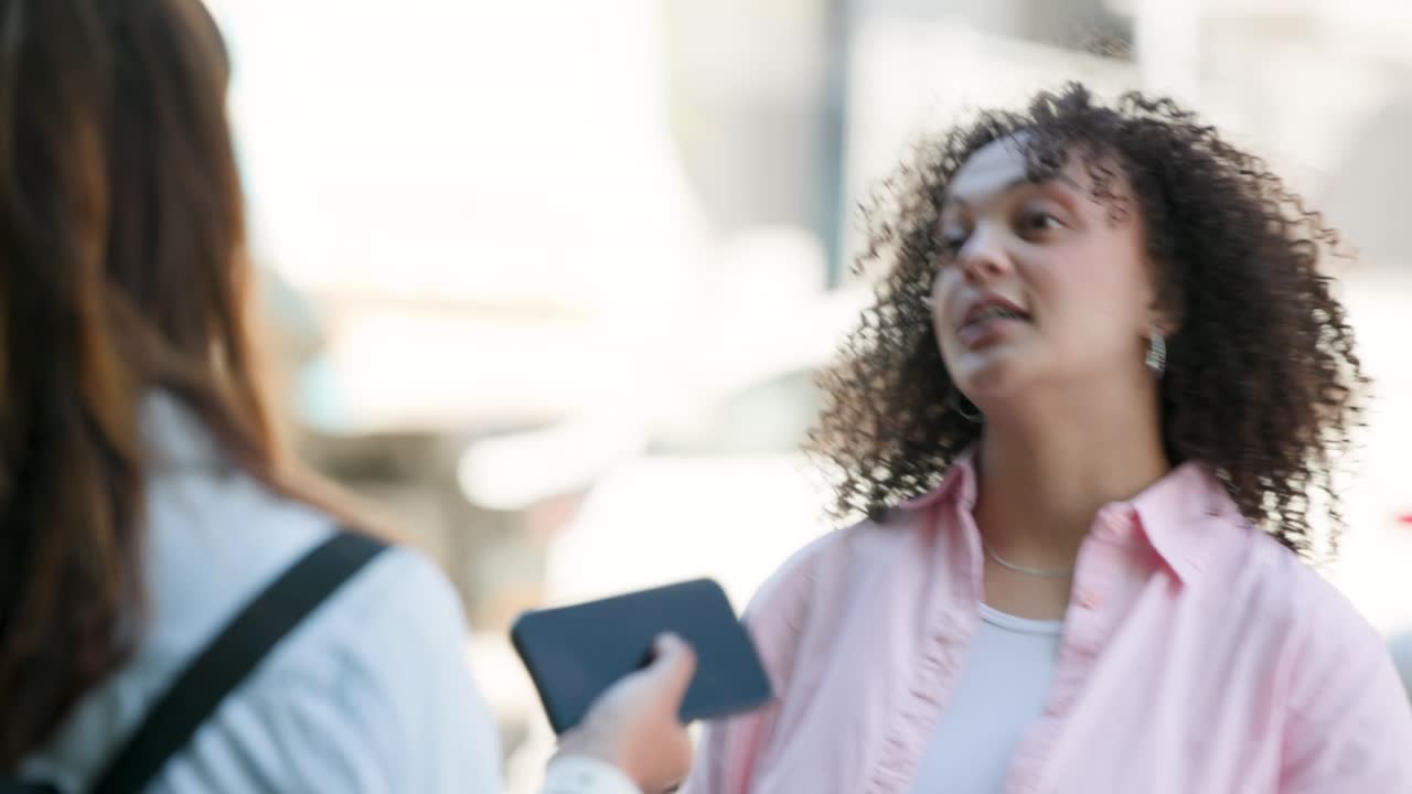 Due donne afroamericane che parlano sul marciapiede, una alza il telefono mostrando una persona riccia che reagisce, mentre un'auto si allontana sfocata.
