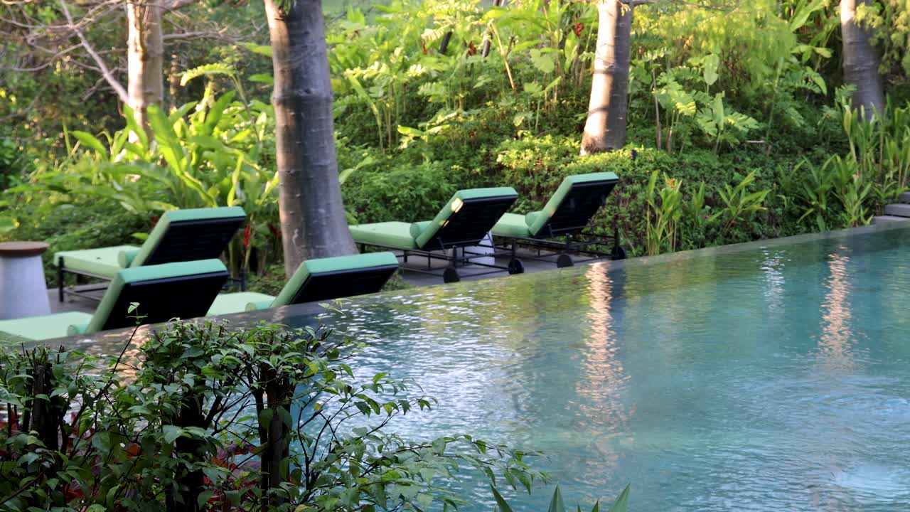 Camera slowly pans across tranquil poolside with green lounge chairs, lush tropical garden, natural lighting