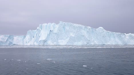 slow motion pan of a large iceberg floating on the ocean, with small ice floes in the foreground under a cloudy sky off the coast of greenland