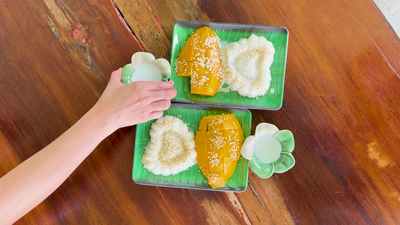 A hand pours coconut milk over mango sticky rice on a wooden table in a bright, tropical setting