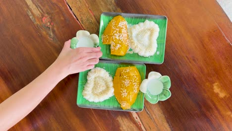 a hand pours coconut milk over mango sticky rice on a wooden table in a bright, tropical setting