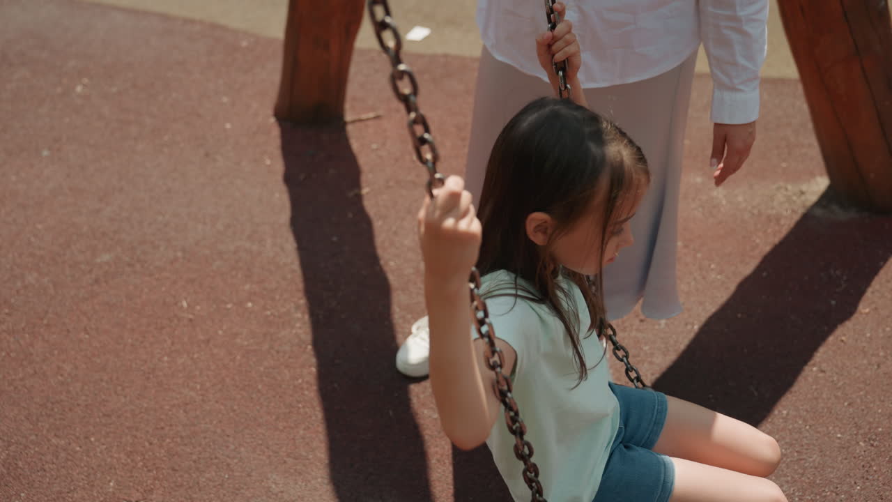 Park swing with mom and daughter playing together on rubberized playground surface, mother standing nearby guiding daughter, warm sunlight casting long shadows, casual outfit with sneakers and skirt