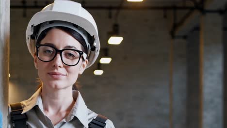 professional female builder wearing safety glasses and hardhat standing confidently inside industrial warehouse, radiating expertise and career success