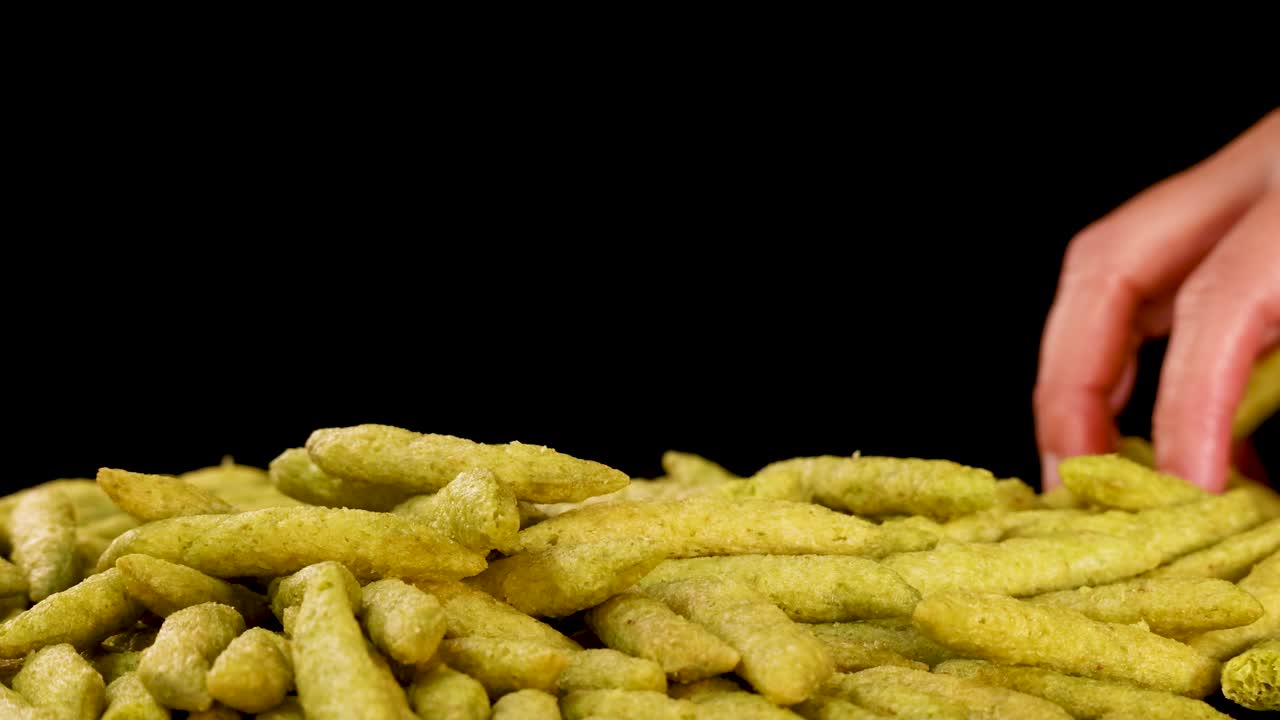 Human hand selects puffed green pea snack sticks from pile, brightly lit, black background, close-up