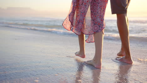 Una-Pareja-Birracial-Camina-Descalza-Por-La-Playa-Al-Atardecer,-Una-Mujer-Vistiendo-Un-Vestido-Estampado,-Con-Copia-Sp