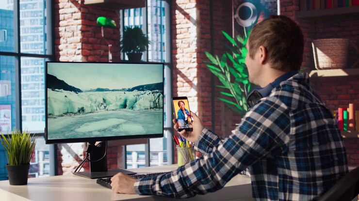Homem usando o telefone, com o monitor do PC ocioso à sua frente exibindo um protetor de tela.