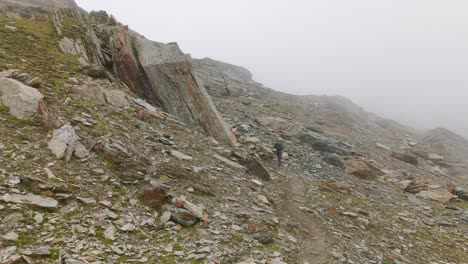 Aerial-forward-view-of-hiker-walking-in-rugged-rocky-mountain-landscape-shrouded-in-fog-in-Valmalenco,-Italy