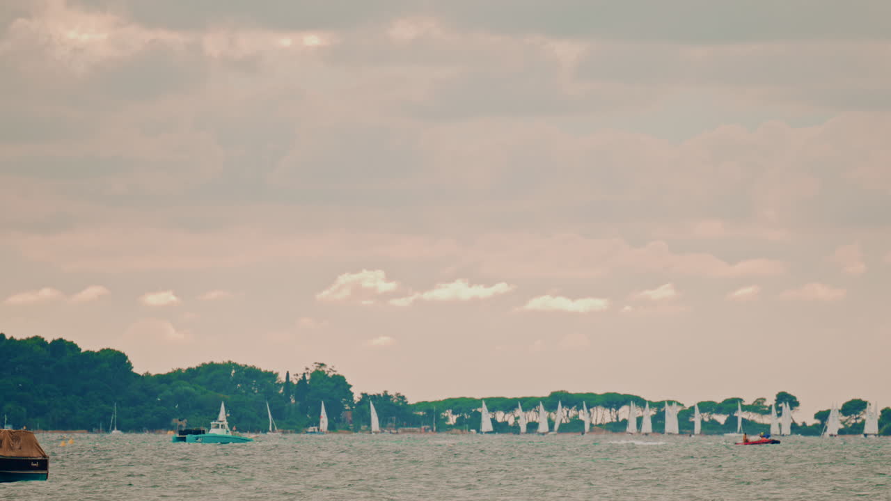 Dozens of small sailboats race across the distant horizon under a cloudy sky, seen from the open sea