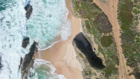waves crash on golden sand by cliffs, praia de samoqueira, alentejo, portugal