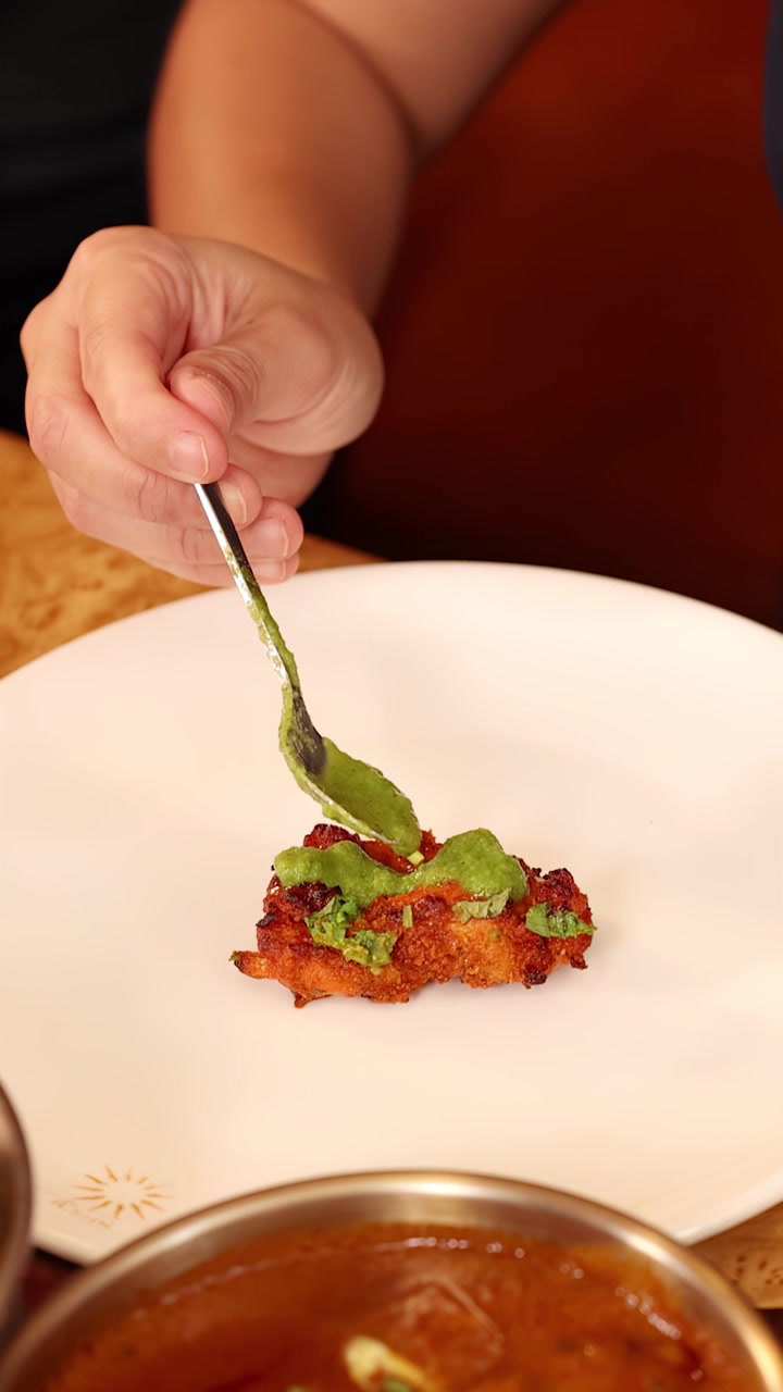 Close-up shot of hand drizzling green sauce onto chicken tikka on white plate.