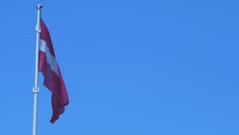 gran bandera letona ondeando lentamente en el cielo azul en un día soleado, avión volando a través del fondo, plano medio