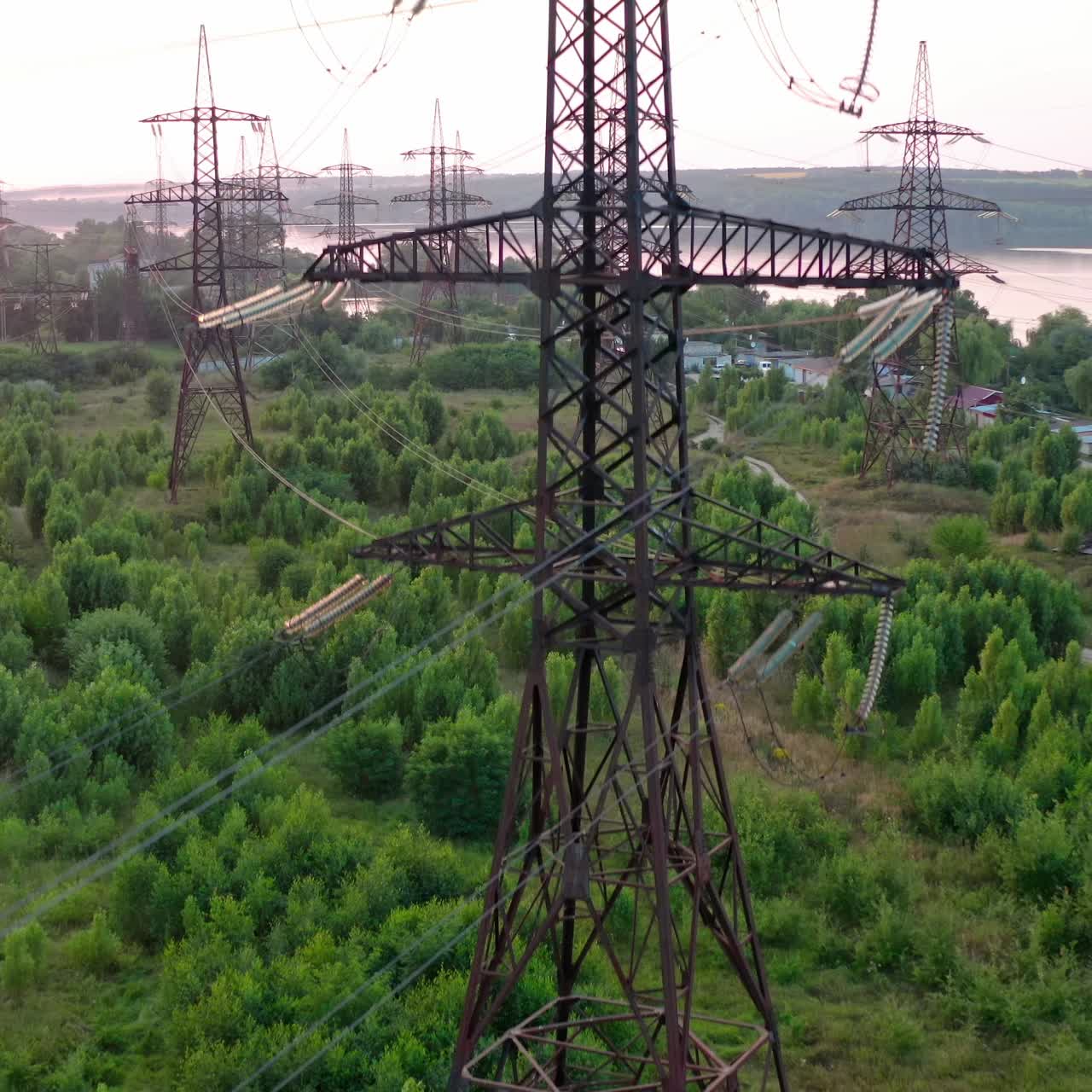Structure of high-voltage lines outside the city at sunset. Trees and river. Camera motion back. Aerial view