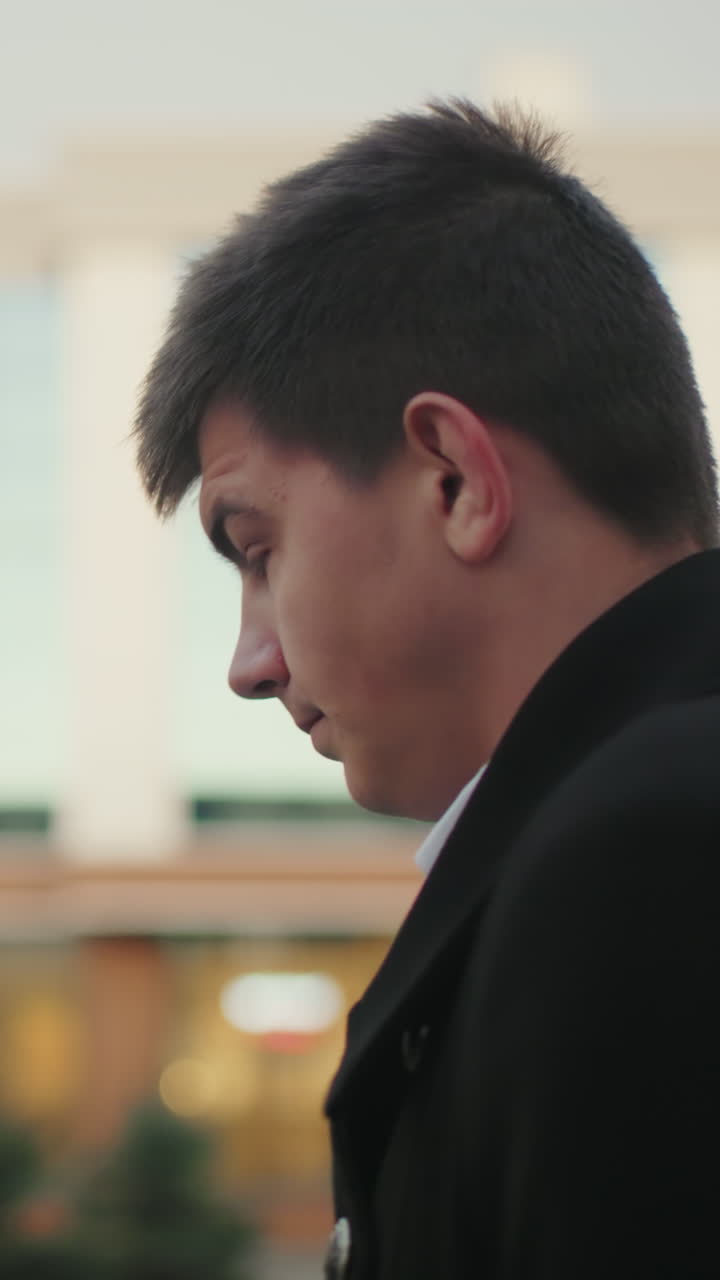 검은 옷을 입은 남자가 밖으로 나가서 시간을 숙고하며, 배경에는 유리 건물이 있으며 부드러운 빛의 반임, 도시 환경에서 세련되고 자신감있는 개인이 있습니다.