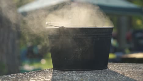 Dampfender-Wascheimer-Aus-Metall-Auf-Einem-Picknicktisch-Aus-Beton