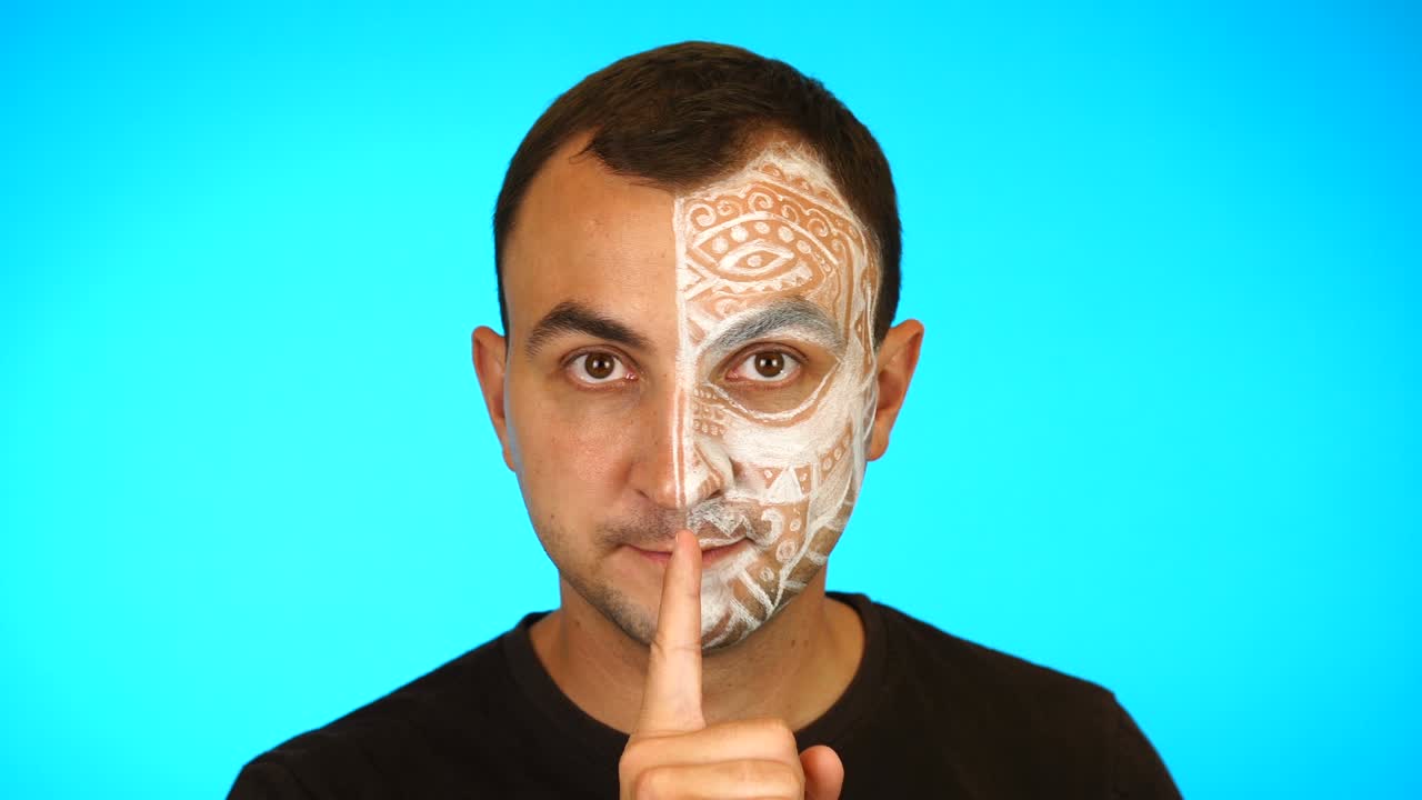 hombre con cara pintada está pidiendo silencio poniendo el dedo en los labios contra un fondo azul