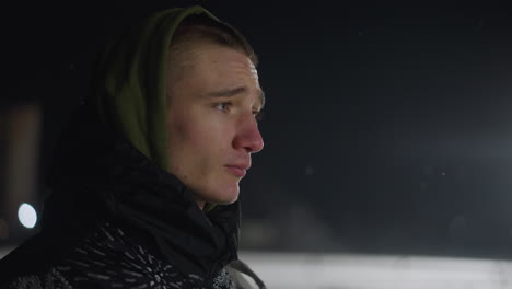 close up of young man in winter hooded jacket staring thoughtfully into distance under intense stadium lights at night during cold weather, with snow visible in blurred background and flurries in air