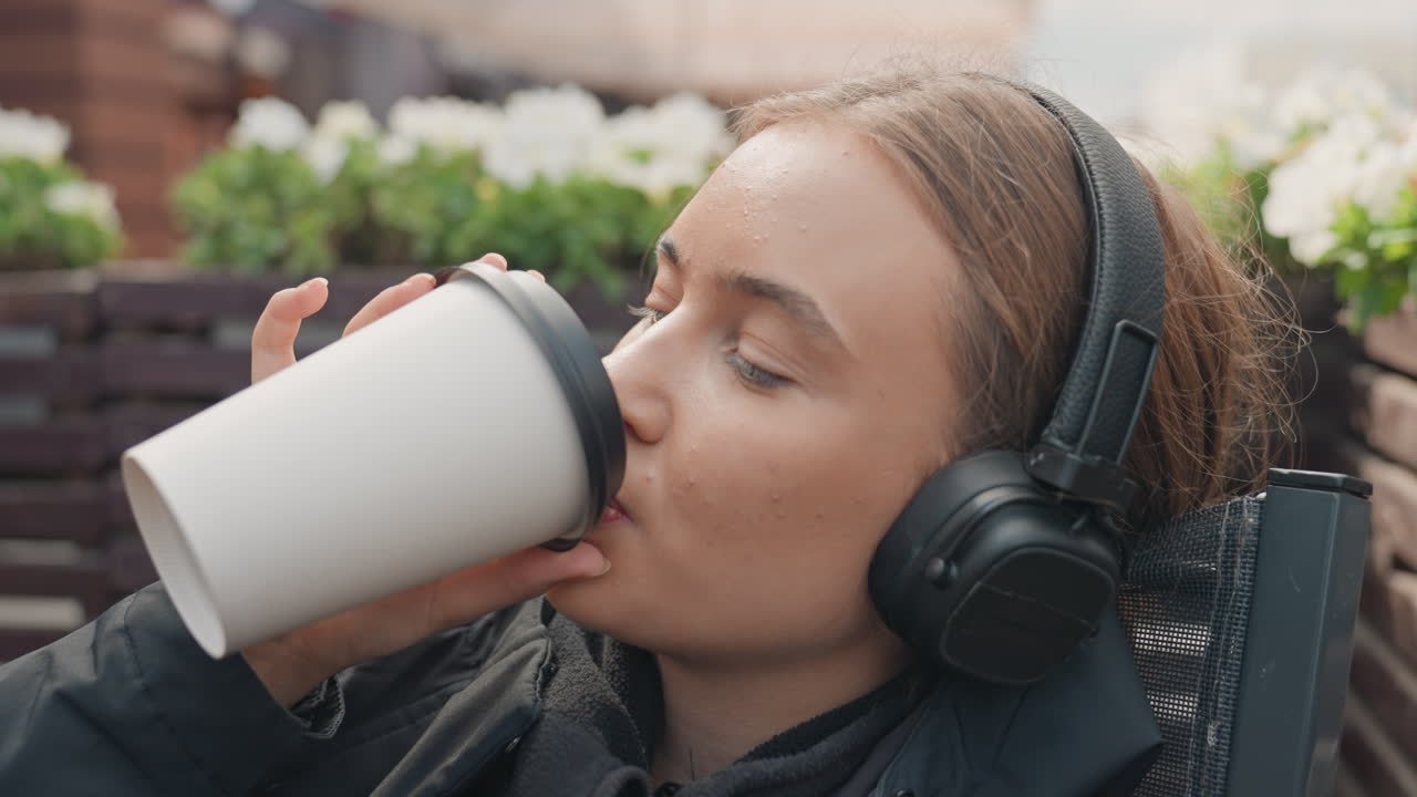 ung kvinna på terrassen som lyssnar på musik och smuttar på kaffe från en take away-mugg, bär overear-hörlurar och mörk jacka, avslappnat leende och daggfrisk hy, närbildsporträtt och breda bilder med trä-