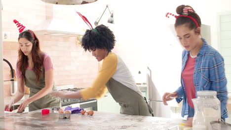 women friends baking christmas cookies together in kitchen, wearing festive headbands