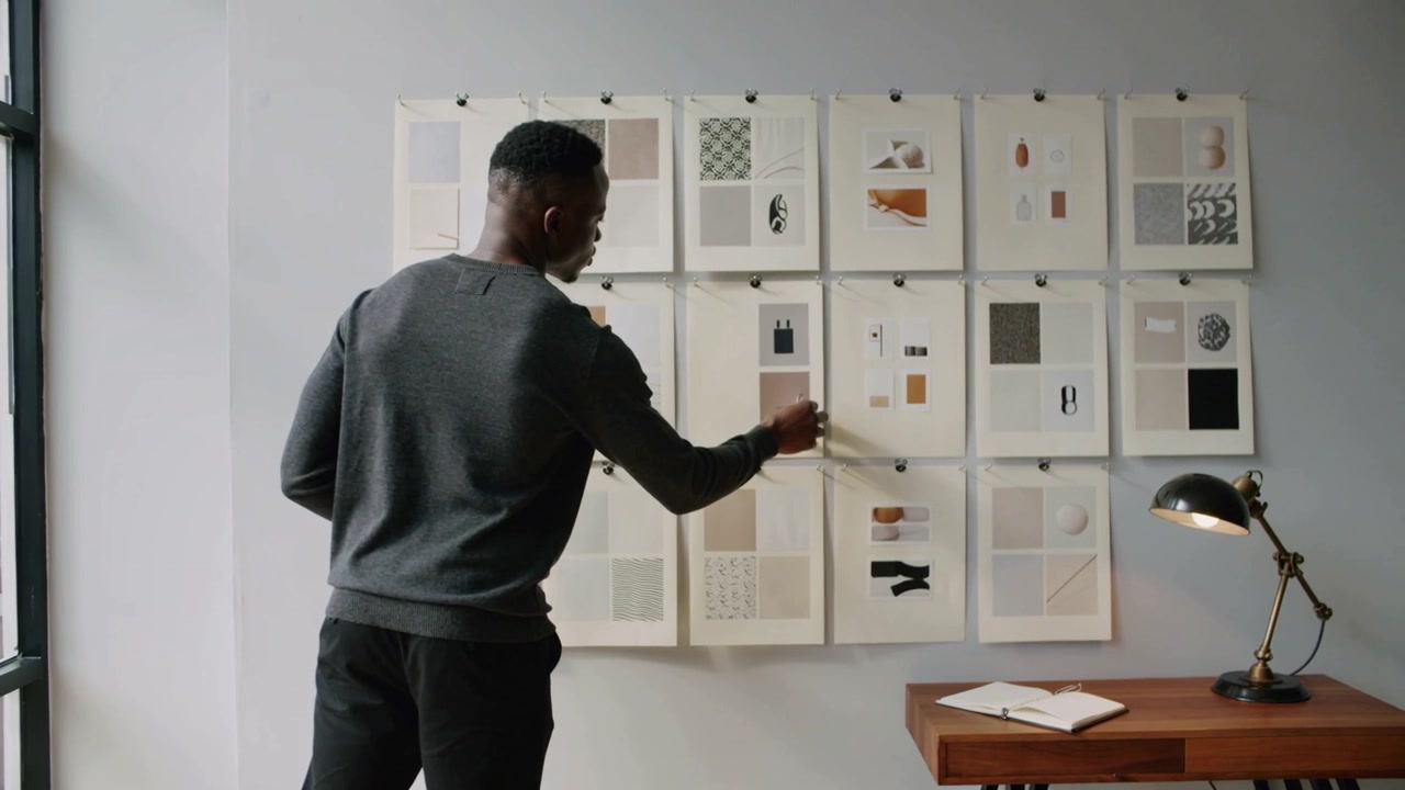 Un homme en pull foncé, entrant par la gauche, ajuste des tirages épinglés avec des pinces dans un studio, finalisant la mise en page.