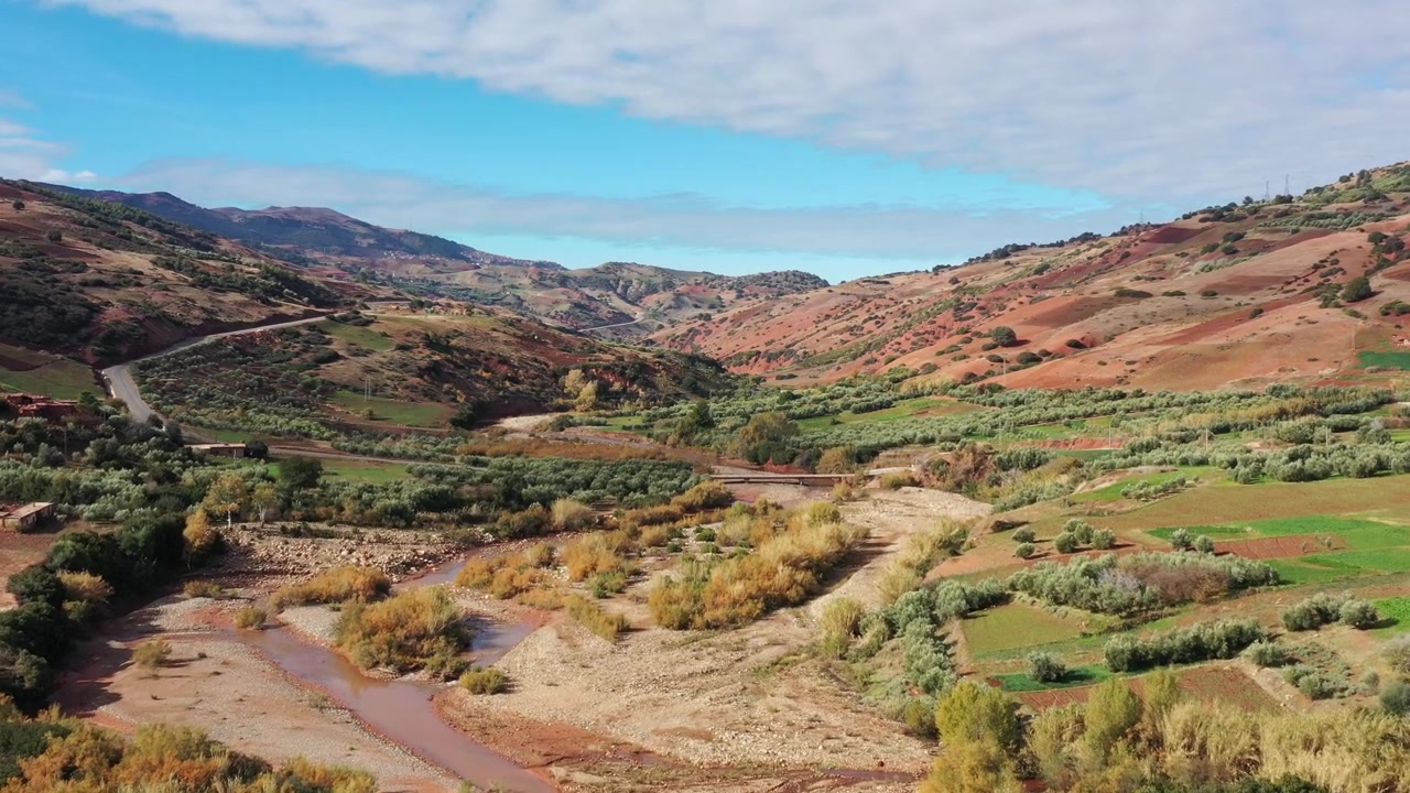 Valle montañoso escénico marroquí con río caudaloso, vegetación verde y colinas onduladas bajo un cielo azul claro. Paisaje natural cinematográfico ideal para proyectos de viajes y documentales.