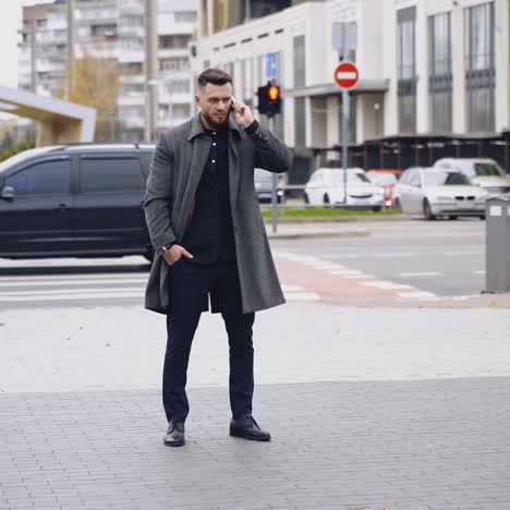 street style. man in an elegant black suit and gray coat. handsome young caucasian man crossing a central road talking on a phone