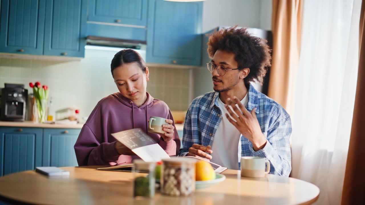 Coppia mista che fa colazione in una cucina accogliente, la donna beve caffè leggendo.