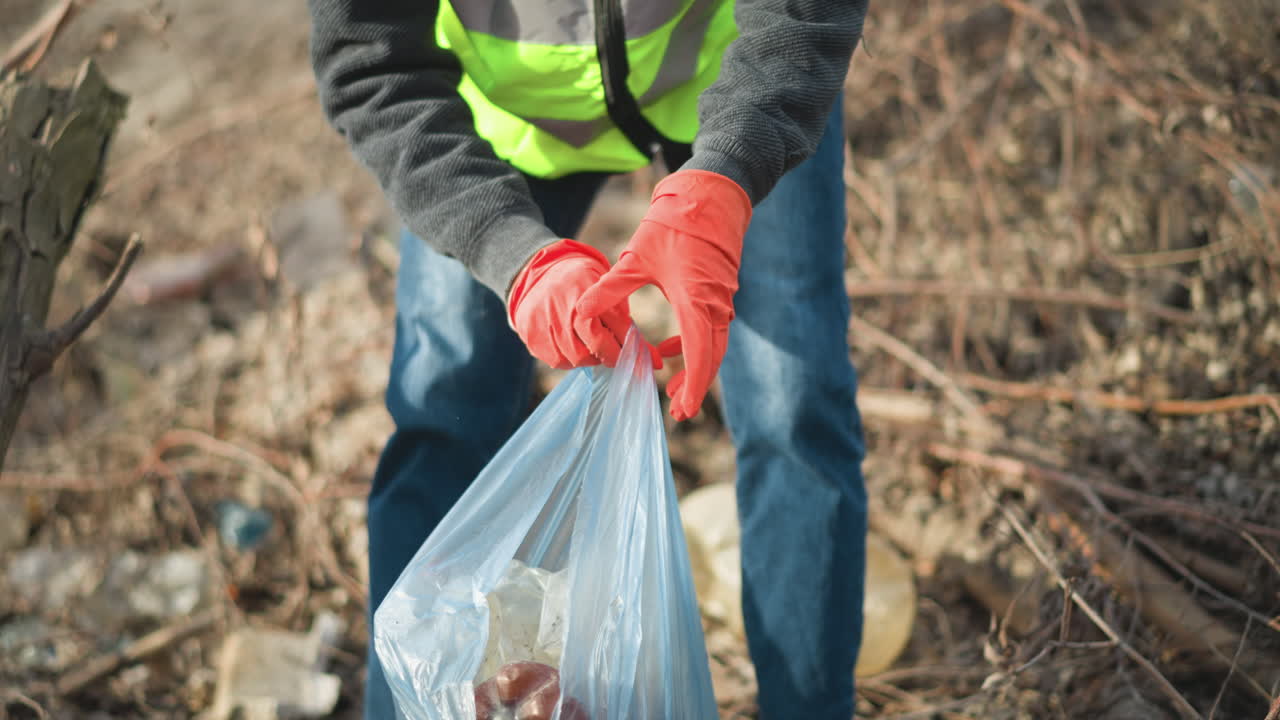 Volunteer wearing orange gloves holding blue plastic bag filled with collected trash, including cans and bottles, while picking up discarded glass bottle during outdoor cleanup to protect environment