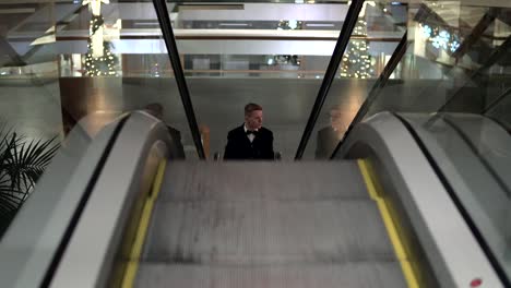 Ein-Junger-Mann-In-Einem-Schwarzen-Anzug-Mit-Fliege-Kommt-In-Einem-Einkaufszentrum-Auf-Die-Rolltreppe