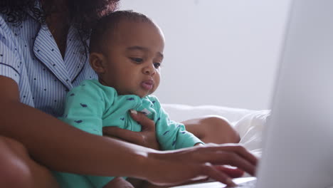 Mother-Wearing-Pyjamas-Cuddling-Baby-Son-In-Bedroom-Whilst-Working-From-Home-On-Laptop