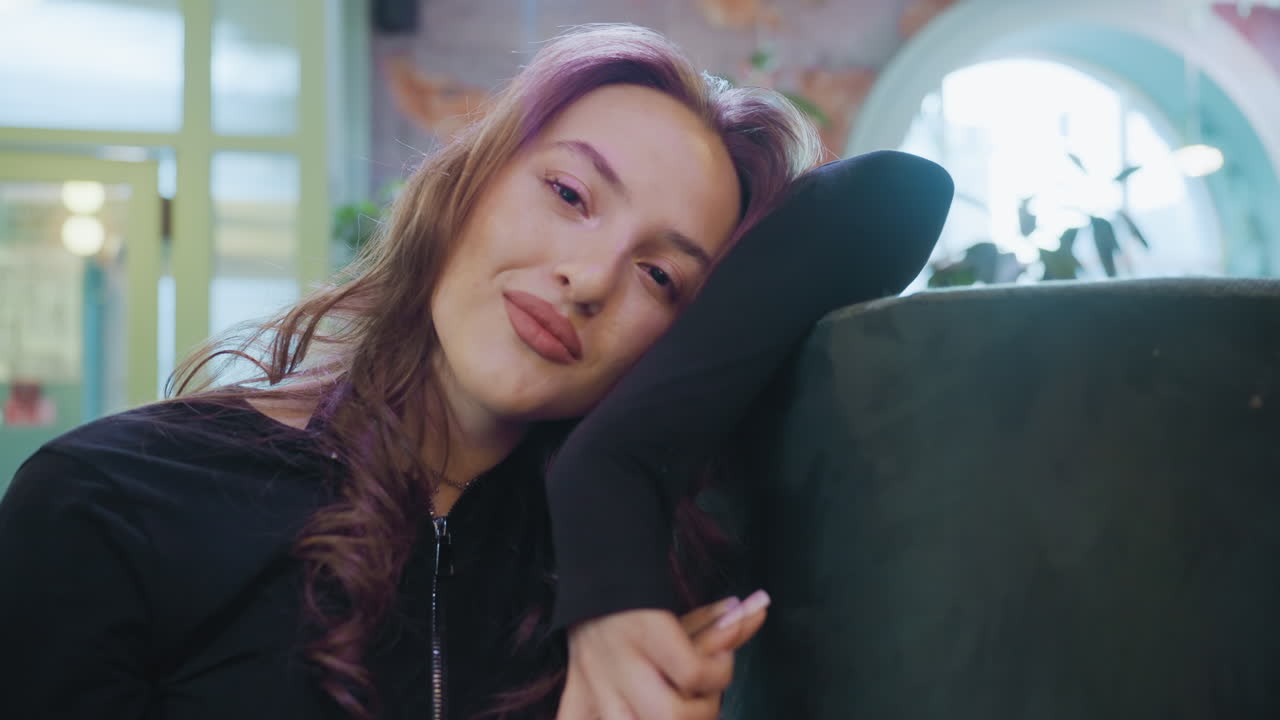 Charming woman with curled hair leans on cushion and looks at camera with gentle smile in softly lit interior space with blurred background, radiating calm confidence and warm personality