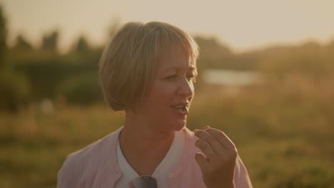 woman eating black grape outdoors under warm sunlight, smiling while looking at someone off-camera, soft focus background features greenery