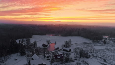 Vista-Aérea-Del-Paisaje-De-La-Puesta-De-Sol-De-Invierno-Rojo,-Naranja-Y-Rosa,-Casas-De-Campo-Con-Un-Lago-En-Los-Bosques